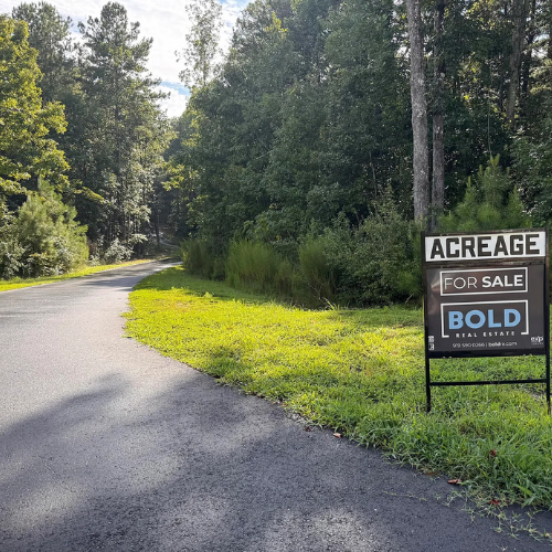 Untitled design 5 - BOLD Construction and Renovations Paved country road curving through a forested area with a for-sale sign at the roadside (acreage for sale).